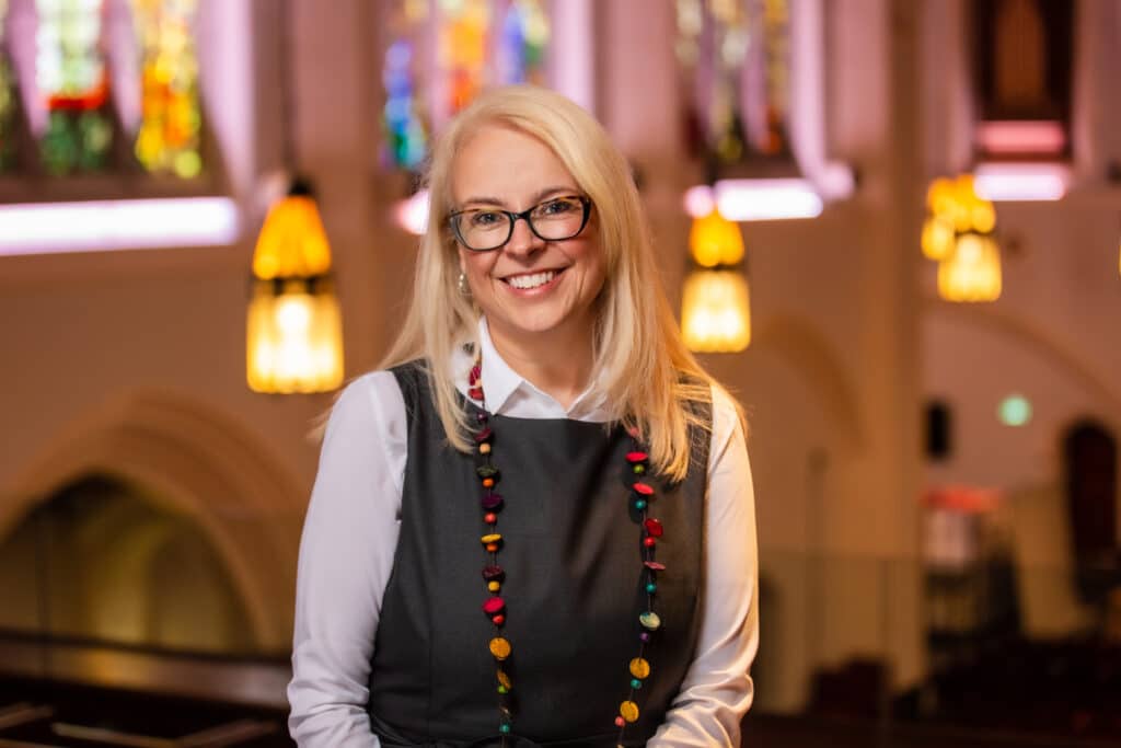 smiling woman with blonde hair in grey dress with stainglass windows background